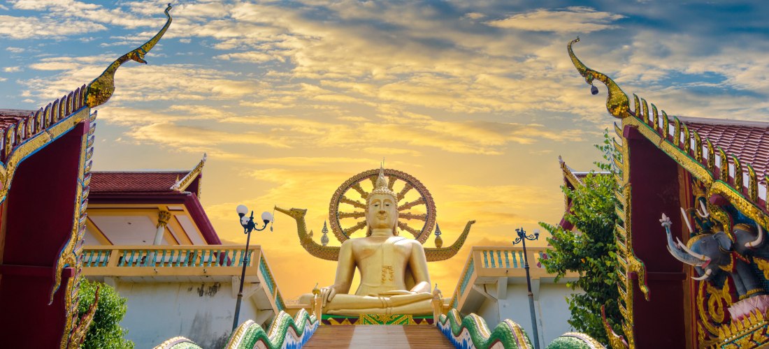  Das Bild zeigt eine monumentale goldene Buddha-Statue, die auf einer Plattform über einer langen, breiten Treppe thront. Die Treppe ist bunt gestaltet, mit wellenartigen grünen und blauen Elementen am Rand, die an Nagas – mythische Schlangenwesen – erinn