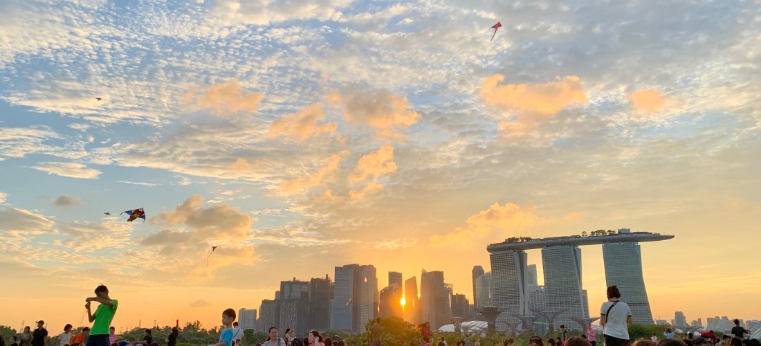 Menschen genießen ein Picknick auf einer großen Wiese im Marina Barrage Park in Singapur. Die Skyline mit dem markanten Marina Bay Sands Hotel ist im Hintergrund sichtbar. Der Himmel ist bunt gefärbt durch die untergehende Sonne, am Himmel fliegen mehrere