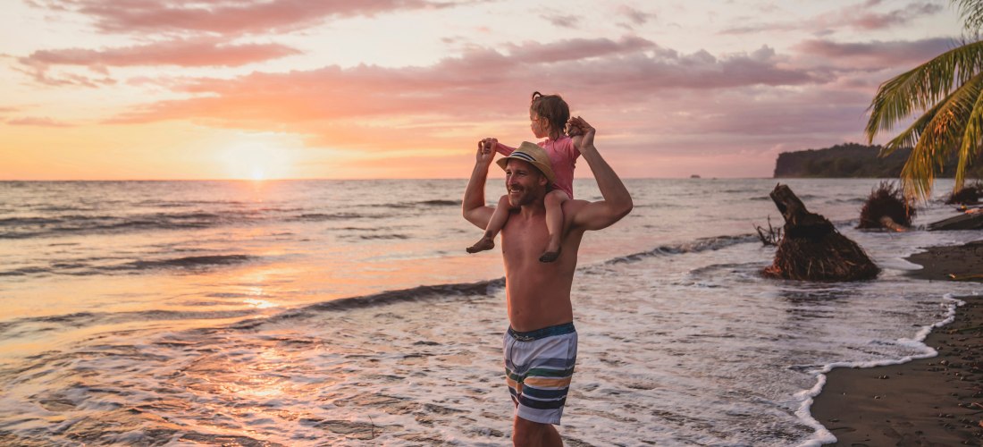 Vater trägt seine Tochter auf den Schultern beim Spaziergang am Strand während eines farbenfrohen Sonnenuntergangs