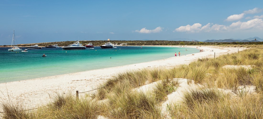 Breiter Sandstrand der Playa de Ses Illetes auf Formentera mit türkisfarbenem Meer. Menschen liegen mit Sonnenschirmen am Strand, während Segelboote im Hintergrund auf dem Wasser ankern.
