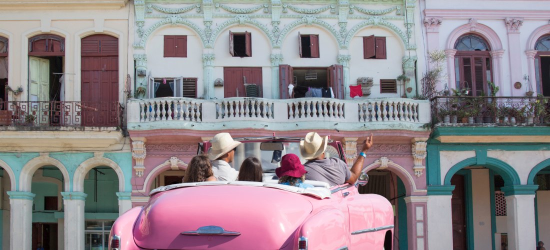 Pinkfarbener Oldtimer mit vier Personen fährt durch eine Straße mit bunten Kolonialgebäuden