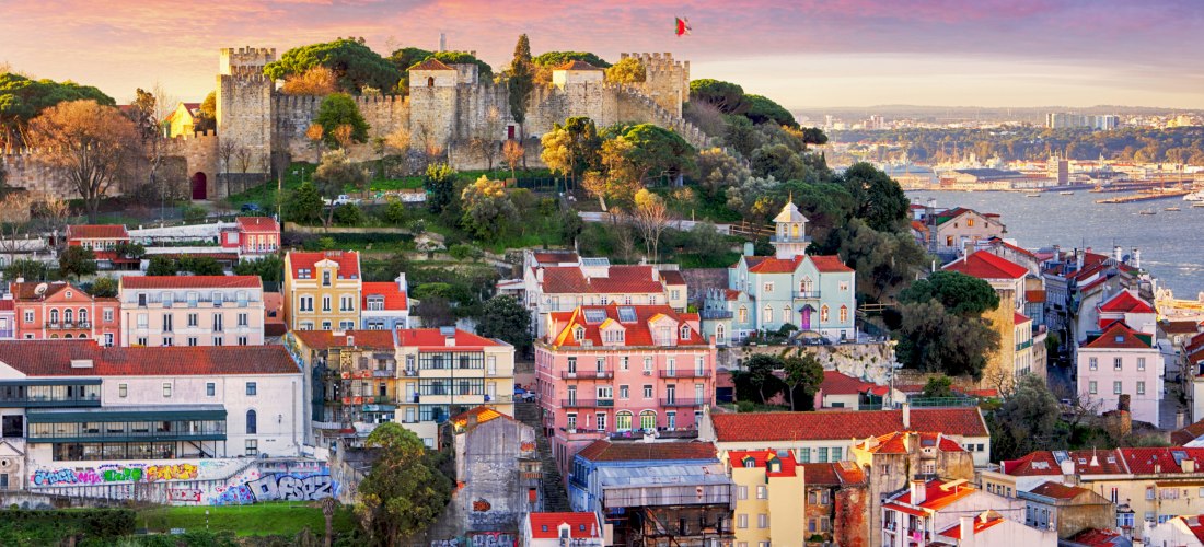 Blick über Lissabon bei Sonnenuntergang: vorn bunte Häuser mit roten Ziegeldächern, darüber die Burg Castelo de São Jorge auf einem grünen Hügel; rechts der Tejo mit Hafenanlagen.