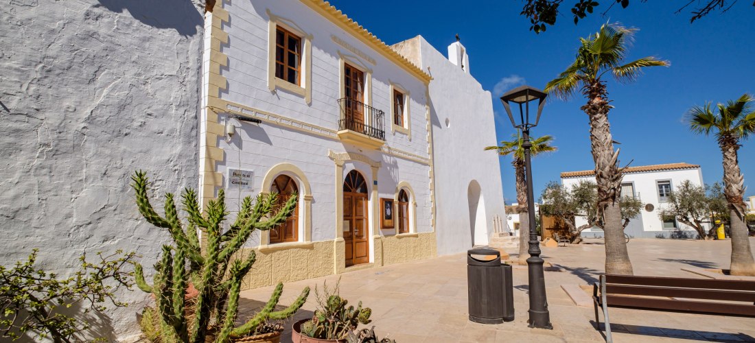 Helle, historische Gebäude ( Städtchen Sant Francesc de Formentera ) mit weißer Fassade und braunen Fensterläden in einem sonnigen Dorfplatz auf Formentera. Im Vordergrund stehen Blumentöpfe mit Kakteen, eine Holzbank und Palmen säumen den Platz. Die Szen