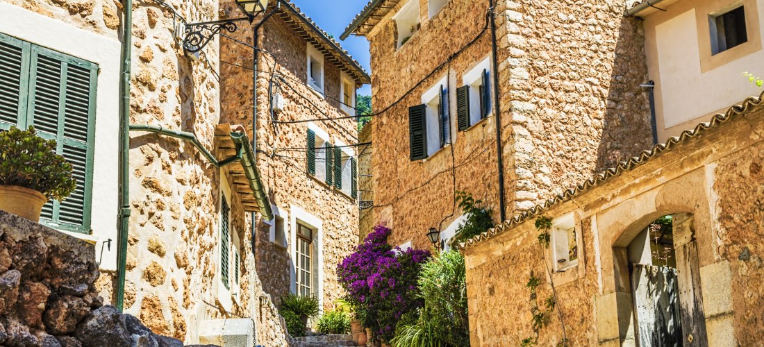 Eine idyllische enge Gasse in einem mallorquinischen Dorf mit traditionellen Sandsteinfassaden und grünen Fensterläden. Bunte Blumentöpfe mit Bougainvillea und anderen mediterranen Pflanzen schmücken die Treppenstufen. Im Hintergrund ein wolkenloser, blau