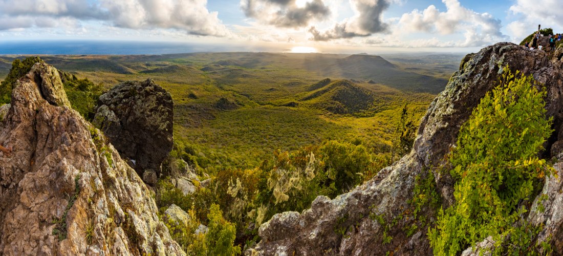Atemberaubender Ausblick vom Gipfel des Christoffelbergs auf Curaçao. Im Vordergrund sind große, zerklüftete Felsen zu sehen, die mit grünem Gestrüpp und Moos bewachsen sind. Dahinter breitet sich eine weite, grüne Hügellandschaft aus, die bis zum Horizon