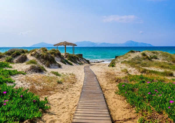 Ein Holzsteg führt durch eine Dünenlandschaft mit grünem Bewuchs und rosa Blüten zu einem türkisblauen Meer. Im Hintergrund sind Berge und eine strohgedeckte Schattenspender-Struktur zu sehen. Der Himmel ist hellblau mit wenigen Wolken.