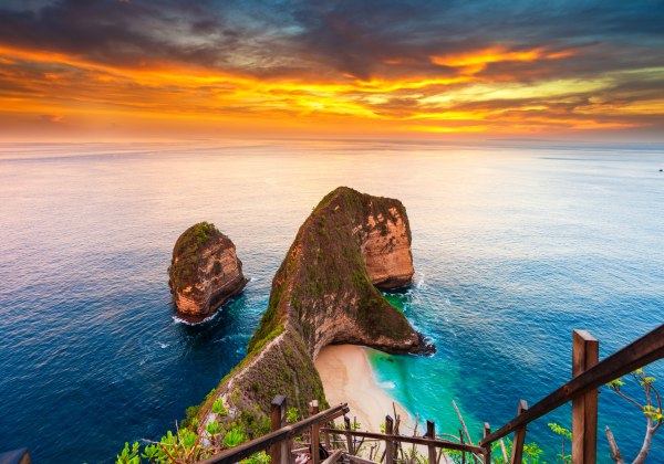 Spektakuläre Aussicht auf den berühmten Kelingking Beach auf der indonesischen Insel Nusa Penida bei Sonnenuntergang. Im Vordergrund führt ein schmaler Holzpfad mit Geländer hinab zur sichelförmigen Bucht mit weißem Sand und türkisfarbenem Wasser. Die mar