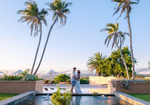 Ein Paar steht in romantischer Pose an einem kleinen Teich, eingerahmt von Palmen, mit Blick auf ein luxuriöses Resort im Hintergrund. Die Szene spielt sich bei klarem Himmel und weichem Abendlicht ab – perfekte Urlaubsstimmung.