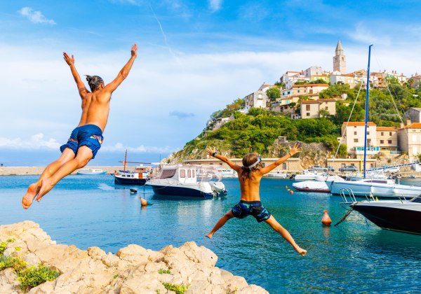 Ein Vater und sein Kind springen gleichzeitig von einem Felsen ins Meer. Im Hintergrund liegt ein kleiner Küstenort mit bunten Häusern am Hang, Booten im Wasser und einem Hafen.