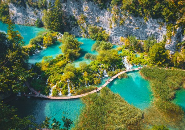 Holzsteg führt durch türkisfarbene Seenlandschaft mit Wasserfällen, üppiger Vegetation und steilen Felswänden im Plitvicer Nationalpark in Kroatien