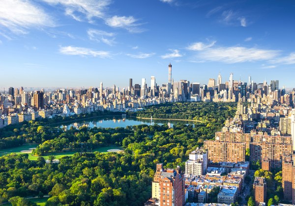 Panoramaaufnahme von New York City mit dem Central Park im Vordergrund. Der grüne Park bildet einen starken Kontrast zur umliegenden Skyline aus hohen, modernen Gebäuden. Der Himmel ist blau mit wenigen Wolken.