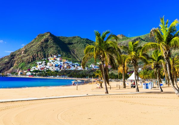 Sandstrand mit Palmen vor einem türkisblauen Meer, im Hintergrund bunte Häuser am Berghang unter klarem, blauem Himmel