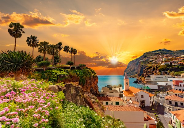 Blick auf eine Küstenstadt auf Madeira bei Sonnenuntergang. Im Vordergrund blühende Pflanzen und Palmen, dahinter Häuser mit roten Dächern auf einem Felsen über dem Meer. Die Sonne strahlt durch orange-goldene Wolken in den Atlantik.