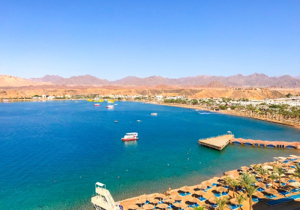 Blick auf eine weitläufige Badebucht mit türkisblauem Wasser, gesäumt von Palmen und Sonnenschirmen. Am Ufer führt eine Promenade mit Steg ins Meer. Mehrere Ausflugsboote und Yachten liegen auf dem Wasser. Im Hintergrund sind Wüstenberge und eine flache S