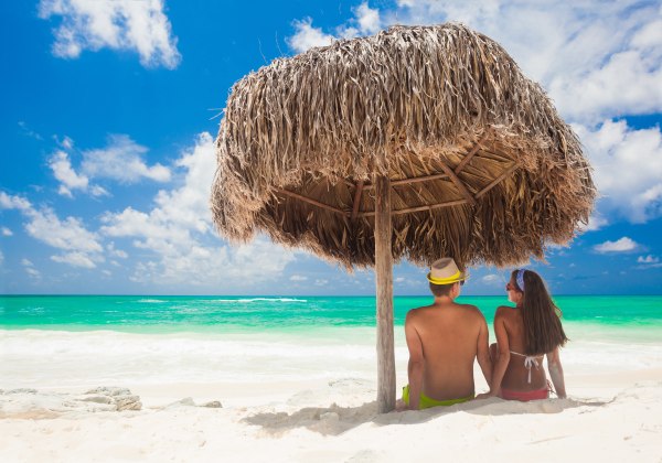 Paar sitzt unter einer Strohhütte am weißen Sandstrand und schaut auf das türkisfarbene Meer