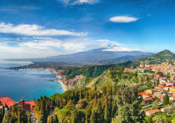 Panoramablick auf die sizilianische Küstenstadt Taormina, die sich terrassenförmig an einen grünen Berghang schmiegt. Rechts im Bild die farbenfrohen Gebäude der Altstadt, mittig der grüne Bergrücken, im Hintergrund der schneebedeckte Vulkan Ätna. Links d