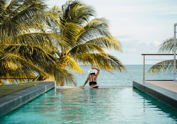Infinity-Pool mit einer Frau in einem weißen Hut, die auf das Meer blickt, umgeben von Palmen und tropischer Vegetation