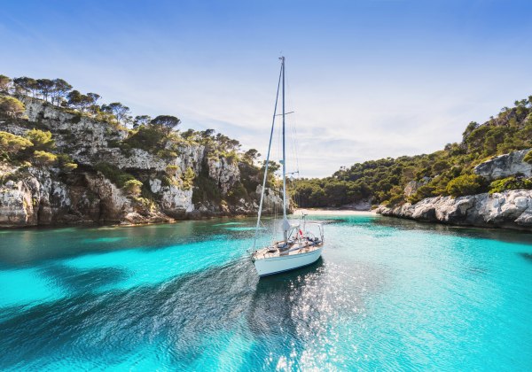 Ein einsames Segelboot gleitet durch kristallklares, türkisfarbenes Wasser in einer malerischen Bucht. Umgeben ist die Szene von felsigen Klippen und üppigem Pinienwald. Die Sonne spiegelt sich glitzernd auf dem Wasser, der Himmel ist strahlend blau – ein