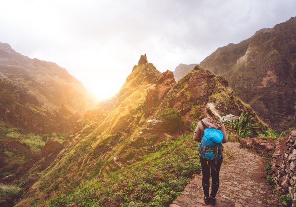 Eine Wanderin mit blauem Rucksack steht auf einem schmalen, gepflasterten Bergweg auf den Kapverden und blickt in ein tiefes, grünes Tal. Um sie herum ragen steile, teils bewachsene Felswände in die Höhe. Die Sonne scheint zwischen den Bergen hervor und t