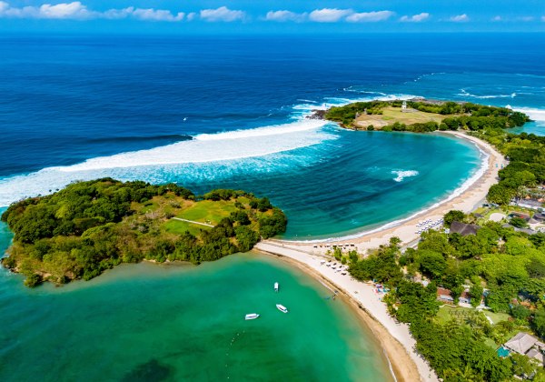 Luftaufnahme einer türkisblauen Bucht mit zwei halbmondförmigen Stränden und grün bewachsenen Inseln; kleinere Boote im ruhigen Wasser, Brandung und offenes Meer im Hintergrund.