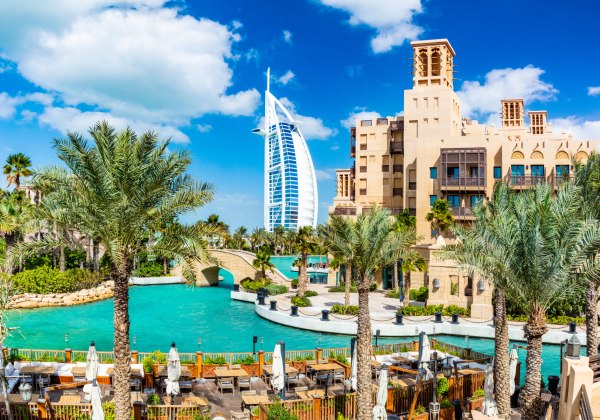 Panoramaaufnahme eines luxuriösen Resorts in Dubai mit Wasserkanälen, Palmen und traditionellen Gebäuden im arabischen Stil. Im Hintergrund ragt das berühmte Hotel Burj Al Arab mit seiner segelförmigen Architektur in den Himmel. Im Vordergrund befinden si