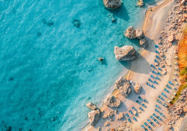Luftaufnahme eines Strandes mit türkisblauem Wasser, Felsen und geordneten Reihen von Sonnenliegen im Sand