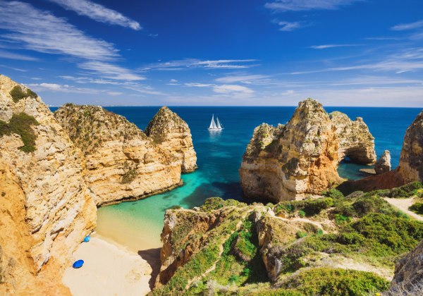 Steile Felsklippen an der Algarve mit kleinen Sandbuchten und türkisblauem Wasser. Im Hintergrund segelt ein weißes Boot auf dem offenen Meer unter blauem Himmel mit leichten Wolken.