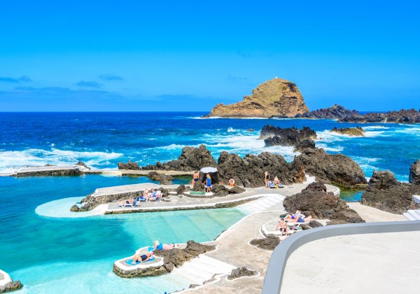Natürliche Lavapools in Porto Moniz auf Madeira mit kristallklarem, türkisfarbenem Wasser, umgeben von schwarzen Vulkanfelsen. Menschen entspannen auf Sonnenliegen unter blauem Himmel mit Blick auf das offene Meer.