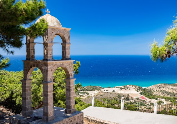 Ein steinerner Glockenturm mit Kreuz steht auf einem Hügel über dem Meer. Im Hintergrund erstreckt sich die türkisblaue Ägäis bis zum Horizont. Umgeben von Pinien vermittelt die Szene Ruhe und mediterrane Atmosphäre.