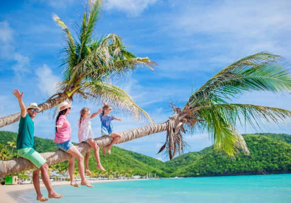 Fröhliche Familie sitzt auf einer geneigten Palme an einem tropischen Strand mit kristallklarem Wasser und grünen Hügeln im Hintergrund
