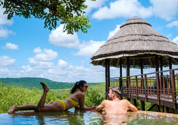 Ein Paar entspannt gemeinsam in einem Infinity-Pool mit Blick ins grüne Buschland. Die Frau liegt auf dem Bauch, der Mann lehnt am Beckenrand. Im Hintergrund steht eine Hütte mit Strohdach, darüber ein blauer Himmel mit weißen Wolken.