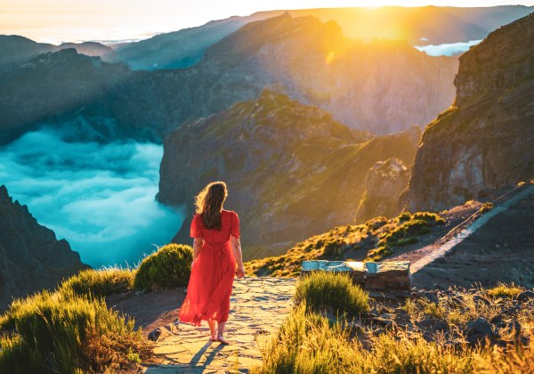 Frau im roten Kleid steht barfuß auf einem Bergpfad auf Madeira und blickt in ein dramatisches Tal, in dem sich Wolken unter ihr sammeln. Die Szene ist in goldenes Morgenlicht getaucht, umgeben von steilen Felswänden und grünen Berghängen