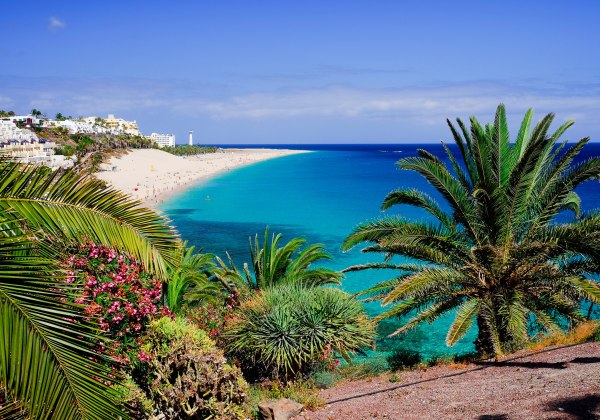 Blick auf einen langen, hellen Sandstrand mit türkisblauem Meer, gesäumt von Palmen, tropischen Pflanzen und Hotelanlagen. Im Hintergrund ein Leuchtturm am Horizont.