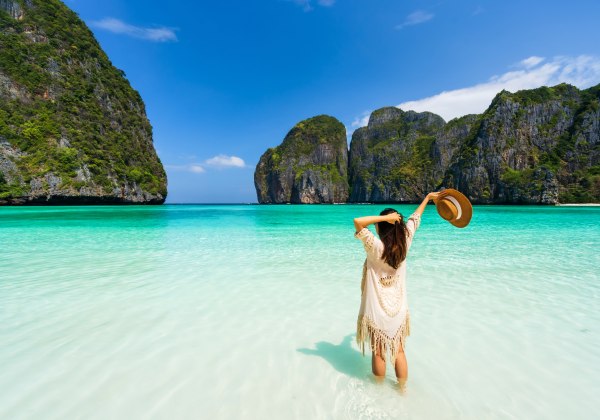 Das Bild zeigt eine Frau, die barfuß im flachen, kristallklaren Wasser der berühmten Maya Bay auf Ko Phi Phi Leh steht. Sie trägt einen cremefarbenen, offenen Strandponcho mit Fransen und hält ihren Sonnenhut in einer Hand, während sie mit der anderen dur