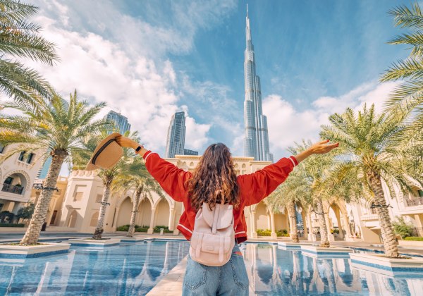Eine junge Frau mit Rucksack steht vor einem Palmenhof und blickt mit ausgestreckten Armen auf den Burj Khalifa in Dubai, den höchsten Wolkenkratzer der Welt. Sie trägt eine rote Jacke und hält einen Sonnenhut in der Hand. Der Himmel ist blau mit einigen 