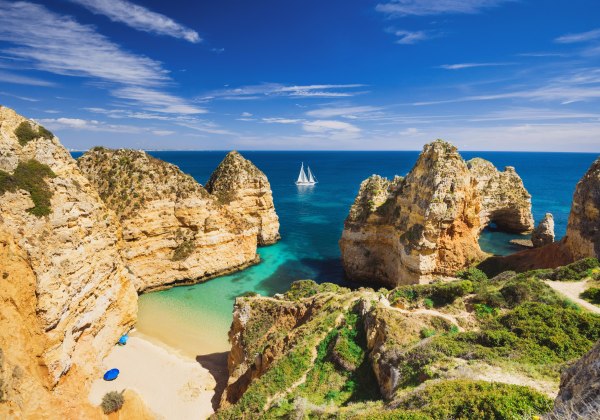 Steile goldene Felsklippen rahmen eine kleine, sandige Bucht mit türkisfarbenem Wasser ein. Auf dem tiefblauen Meer segelt ein weißes Boot, darüber ein klarer Himmel mit dünnen Wolken. Vorn verlaufen schmale Pfade über grün bewachsene Klippen – eine drama
