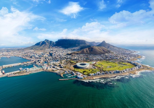 Panoramaaufnahme von Kapstadt, Südafrika, aus der Luft. Im Vordergrund ist das türkisgrüne Wasser des Atlantiks mit Küstenlinie, kleinen Wellen und Hafenanlagen zu sehen. Zentral liegt das große, runde WM-Stadion von 2010, umgeben von grünen Parks und Sta