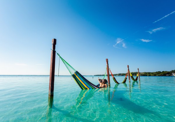 Mann entspannt in einer bunten Hängematte über türkisblauem Wasser, befestigt an Holzpfosten, unter einem klaren blauen Himmel