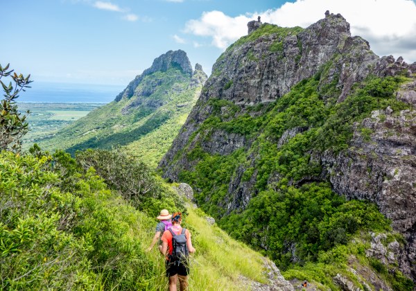 Dieses Bild zeigt eine abenteuerliche Wanderung durch eine atemberaubende Berglandschaft. Zwei Wanderer, ausgestattet mit Rucksäcken und Hüten, bewegen sich vorsichtig auf einem schmalen, grasbewachsenen Pfad entlang eines steilen Hangs. Die Berge sind ei