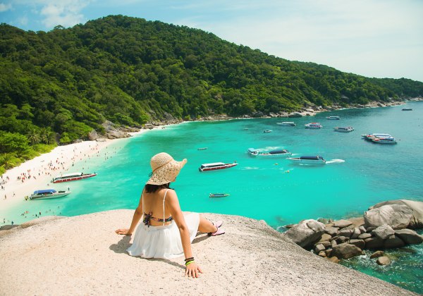 Eine Frau mit Sonnenhut, Bikini und weißem Rock sitzt auf einem Felsen oberhalb einer tropischen Bucht mit türkisfarbenem Wasser. Sie blickt auf einen weißen Sandstrand, an dem viele Menschen baden und entspannen. Im Wasser ankern mehrere Schnellboote und