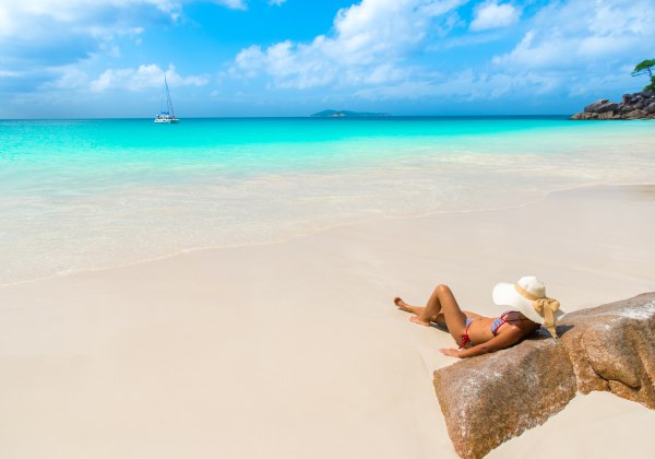 Das Bild zeigt eine Frau, die in einem gestreiften Bikini und mit einem breiten Sonnenhut an einem weißen Sandstrand entspannt. Sie lehnt sich an einen Felsen, während sanfte Wellen das Ufer erreichen. Das Meer leuchtet in verschiedenen Türkistönen, und i