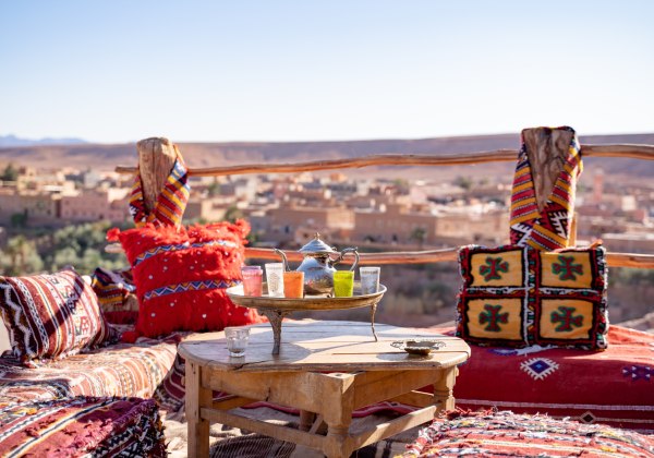 Orientalisch eingerichtete Terrasse mit bunten Kissen, Teetablett und Blick auf eine Wüstenstadt und Berge in der Ferne
