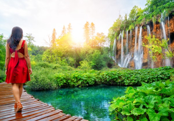 Eine Frau in einem roten Kleid mit offenem Rücken steht auf einem hölzernen Steg, der sich durch üppiges, grünes Naturgebiet schlängelt. Vor ihr liegt ein klarer, türkisfarbener Teich, umgeben von großen, grünen Blättern. Im Hintergrund stürzt eine Reihe 