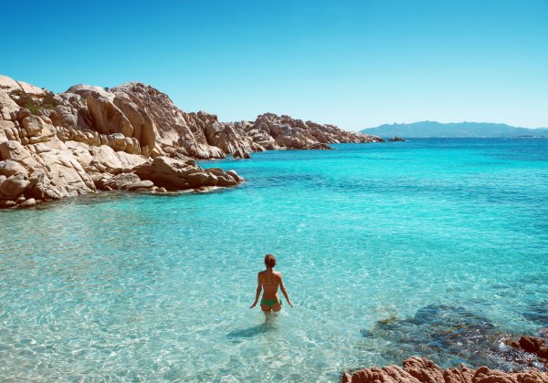 Frau steht im türkisblauen, klaren Wasser vor felsiger Küste unter strahlend blauem Himmel