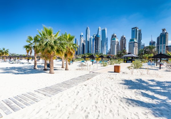 Heller Sandstrand in Dubai mit Palmen, Sonnenliegen und schattigen Pavillons. Im Hintergrund erhebt sich die moderne Skyline von Dubai Marina mit zahlreichen futuristischen Hochhäusern und Wolkenkratzern. Der Himmel ist strahlend blau, die Szene wirkt son