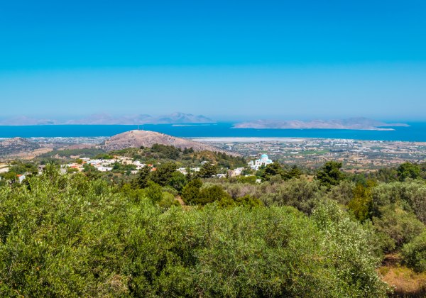 Panoramablick von einem Hügel über die grüne Insel Kos mit ihren verstreuten Häusern, Olivenbäumen und Feldern. Im Hintergrund das tiefblaue Meer mit weiteren Inseln am Horizont unter wolkenlosem Himmel.