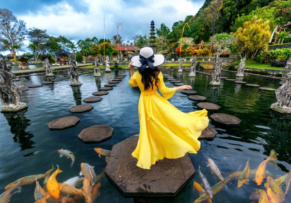 Eine Frau in einem langen, leuchtend gelben Kleid und großem Sonnenhut schreitet über Steine durch ein Wasserbecken im Tirta Gangga Wasserpalast auf Bali. Um sie herum schwimmen farbenfrohe Kois und stehen kunstvolle Steinfiguren. Der Garten ist von tropi