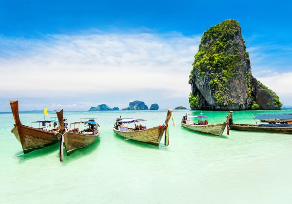 Mehrere Longtail-Boote liegen im flachen, smaragdgrünen Wasser vor einem weißen Sandstrand. Im Hintergrund ragt eine steile, grüne Kalksteininsel aus dem Meer, typisch für die Landschaft um Phuket.