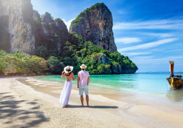 Das Bild zeigt ein Paar in sommerlicher Kleidung, das entspannt an einem tropischen Sandstrand steht. Die Frau trägt ein weißes, luftiges Kleid und einen Sonnenhut, der Mann helle Shorts, ein rosa Shirt und ebenfalls einen Hut. Beide blicken auf das ruhig