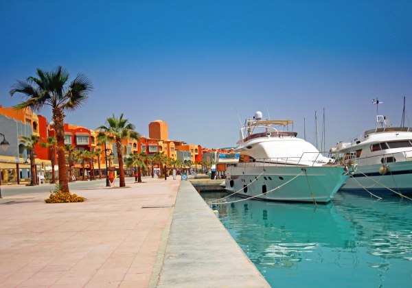 Das Bild zeigt die lebendige Marina von Hurghada am Roten Meer. Rechts im Bild liegen mehrere luxuriöse Yachten und Boote im türkisfarbenen Wasser vertäut. Links verläuft eine breite, gepflegte Promenade mit Palmen, Blumenbeeten und farbenfrohen Gebäuden 